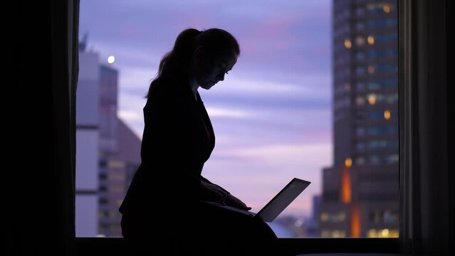 Woman continue work at evening time, sitting at window sill with laptop. Silhouetted shot of business lady, blurred city buildings and sunset sky seen on background