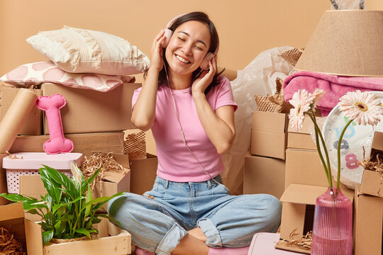 Pleased Asian woman enjoys listening music in stereo headphones sits crossed legs at messy room relocates to new apartment buys new house for living. Happy property owner surrounded by cardboard boxes
