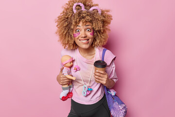 Happy excited curly pregnant woman bites lips poses with takeaway coffee and doll going to become mother soon dressed in casual clothes prepares for giving birth isolated over pink background