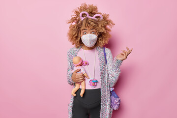 Shocked confused pregnant woman stands unaware wears protection mask stays at home during lockdown and coronavirus spread holds doll carries bag and nipples on string. Motherhood and expectation