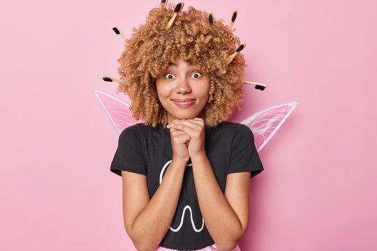 Horizontal shot of surprised curly haired woman pretends being tooth fairy keeps hands under chin has toothbrushes stuck in hair looks with wondered expression isolated over pink background.