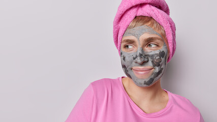 Horizontal shot of thoughtful woman applies beauty clay mask on face focused aside undergoes skin...