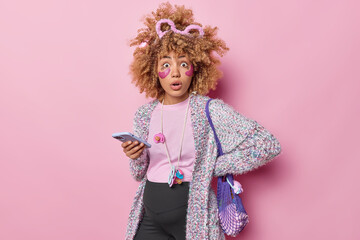 Pregnancy technology and expectation concept. Shocked curly pregnant woman holds smartphone checks notification applies hydrogel beauty pads dressed in casual clothes isolated on pink background