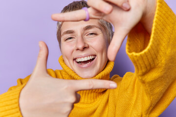 Happy cheerful pretty woman makes frame with fingers smiles toothily captures moment has glad expression dressed in bright yellow jumper isolated over purple background searches perfect angle