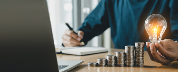Businessman holding and putting lightbulb on coins stack on table for saving energy and saving money concept.