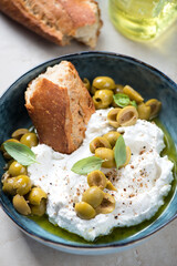 Ricotta cheese served in a bowl with sliced green olives, crusty bread, basil leaves and olive oil, vertical shot, close-up, selective focus