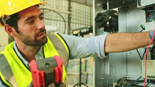 Male electronic technician caucasian checking electrical control system with multimeter or clamp meter at workbench, robotic arm power supply connection, welding cabinet for safety.