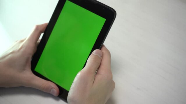 Close-up of a human's hands holding a tablet computer with a green screen layout and making screenshot by swipe, white table background. Top view. Video chat, work, analysis