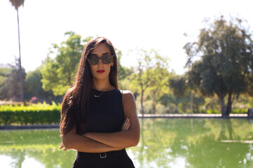 Beautiful brunette Spanish woman with red lipstick. In the background is a lake in the park. The woman is dressed in elegant black clothes and wears sunglasses.