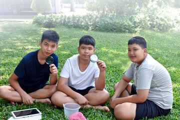 Three Asian studets holding magnifying glasses and other learning devices, tablets, small fishing nets and a small white plastic bin to learn about underwater insects after taking it out of the pond.