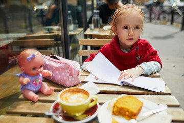 Adorable preschooler girl drawing in cafe while her mother is drinking coffee