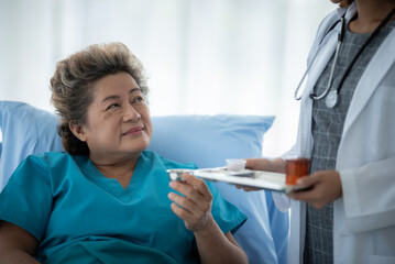 Obraz premium Smiling young doctor and nurse in lab coat with stethoscope giving care and medicinal syrup and treatment to senior woman resting on hospital bed while admitted for treatment against illness