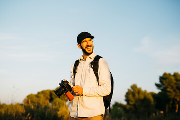 Portrait of a young bearded man, wearing a black cap, with his digital camera at countryside