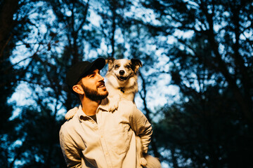 Portrait of a young bearded man, wearing a black cap, carrying a dog on his back at countryside