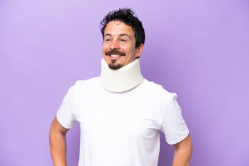 Young caucasian man wearing neck brace isolated on purple background posing with arms at hip and smiling