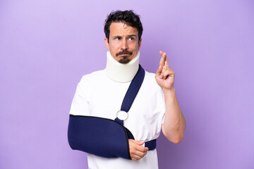 Young caucasian man wearing a sling and neck brace isolated on purple background with fingers crossing and wishing the best