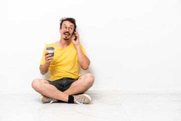 Fototapeta premium Young caucasian man sitting on the floor isolated on white background holding coffee to take away and a mobile