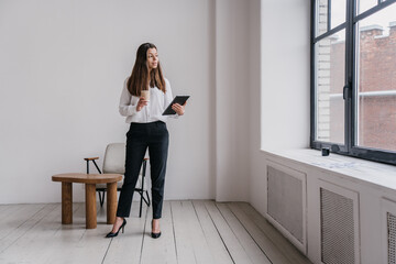 Purposeful hispanic businesswoman in white shirt and black pants standing at office with cup of...
