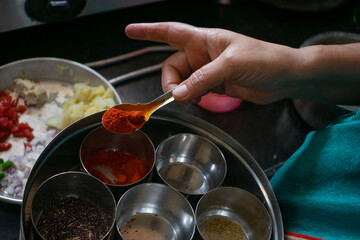 Hand of a woman holding spoon full of chilly powder while cooking food. Main focus is on the hand of the woman. Masala, Garama masala, salt, coriander powder, cummin seeds, turmeric, mustard, potato.