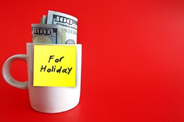 White cup with cash dollars money and yellow stick note with text written FOR HOLIDAY on copy space red background - concept of financial budget planning for coming holiday season