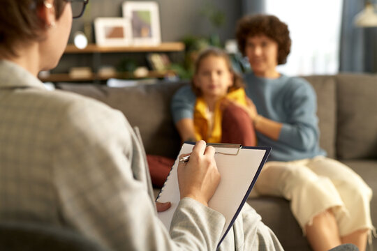Close-up of child psychologist making notes in document while talking to mom and her son during her visit to home
