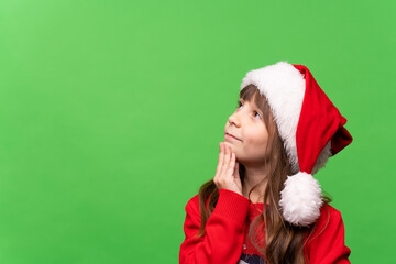A cheerful little girl in a red knitted sweater and a Santa hat looks thoughtfully dreamily up, dreams of a gift, makes wishes. Merry Christmas and Happy New Year 2023