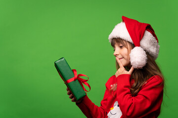 A little girl in a Santa hat holds a Christmas present in her hand and tries to guess the contents of the box. Isolated on green background