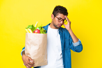Young Brazilian man holding a grocery shopping bag isolated on yellow background with headache