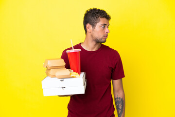 Young brazilian man holding pizzas and burgers isolated background looking to the side