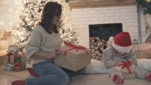 Merry Christmas family holiday gift. Funny little girl opens gift box while sitting on floor near Christmas tree. Mom and daughter unwrap Christmas present. Concept of holiday, celebrations