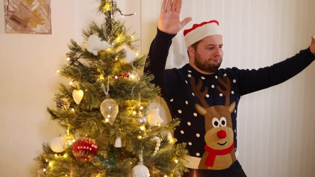 Young happy man with a Rudolf Xmas jumper and a Santa hat dancing to the music while standing near glowing Christmas tree