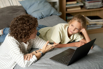 Mother talking to her daughter while they lying on bed and using laptop in bedroom