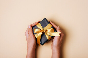 Girl's hands holding black gift box with golden bow on neutral beige background. Christmas, Valentines day, Black friday sale concept. Top view, flat lay