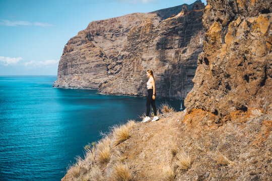 Independent relaxed calm slim girl tourist stands on the edge of a cliff with amazing ocean and sky view. Los Gigantes mountain ranges. Tenerife. Santiago del Teide. Canary Islands, Spain.
