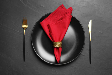 Plate with red fabric napkin, decorative ring and cutlery on black table, flat lay