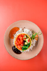 Raw red fish, avocado, seaweed, red caviar, sesame seeds in orange peel with rice and sauces on a red background.