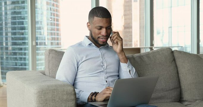Handsome millennial Black business man talking on cellphone in hotel room, holding laptop on lap, typing, using online app for phone call conversation, sitting on couch at panoramic window behind