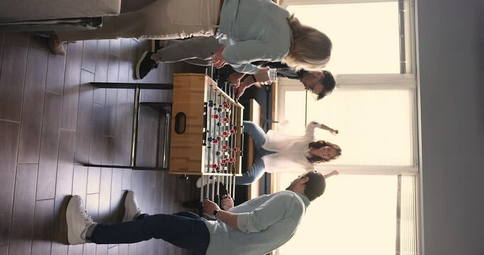 Cheerful employees playing table foosball during break indoors, vertical shot. Workmates cheering, celebrate win, giving high five gesture feel happy enjoy rest at workplace. Friendship, office games