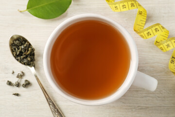 Cup of herbal diet tea and measuring tape on light wooden table, flat lay. Weight loss concept