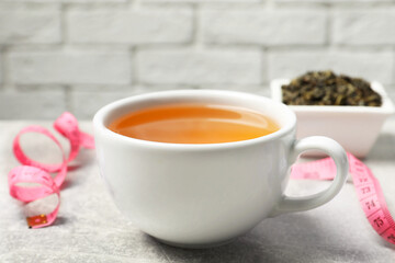 Cup of herbal diet tea and measuring tape on light grey table against brick wall, closeup. Weight loss concept