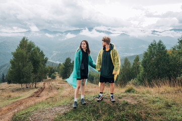 Young beautiful couple in raincoats holding hands standing in the mountains against the background of beautiful views and looking at beautiful scenery during a hike