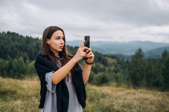 Attractive female tourist hiking in the mountains stands with a smartphone in her hands and takes a photo on the camera with a serious face and looks away on the background of landscape with mountains