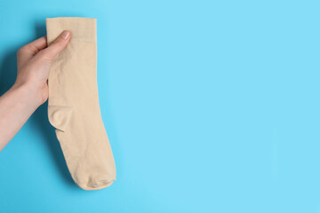Woman holding beige sock on light blue background, closeup. Space for text