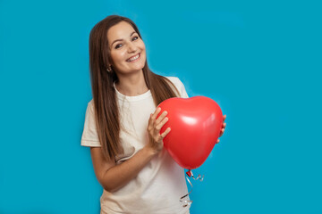 Young beautiful girl smiles happily with a heart-shaped balloon in her hands on a blue background