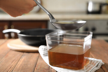 Woman pouring used cooking oil with ladle into container on wooden table in kitchen, closeup