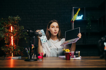 Beautiful woman beauty blogger is recording talk videos with eyeshadow palettes in hands in...