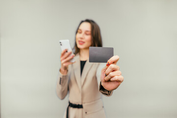 Woman on a beige background with a smartphone in his hands shows the bank card to the camera. Black credit card in the hands of a business woman.