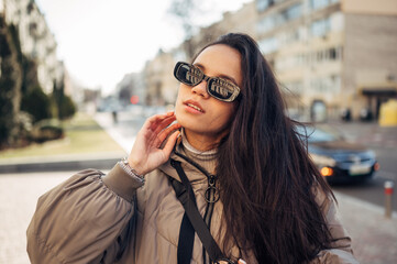 Portrait of a beautiful woman in sunglasses and a warm jacket stands on the street in the autumn day and looks away.