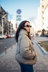Fototapeta premium Portrait of a beautiful woman in warm casual clothes and sunglasses walking down the street on a warm spring day. Vertical