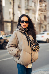 Obraz premium Portrait of a beautiful Hispanic woman in warm clothes and sunglasses standing on the street and looking at the camera with a serious face. Vertical